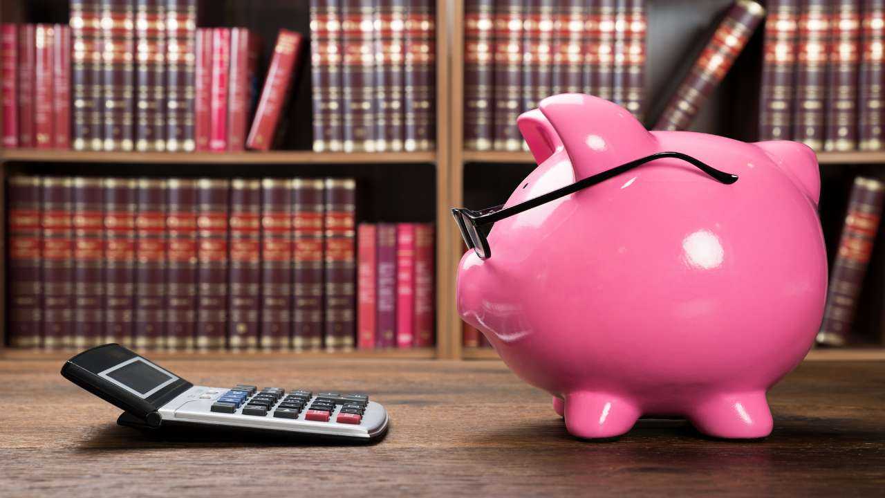 На фоне книг на столе стоит калькулятор и свинка-копилка в очках – важность основ бюджетирования
