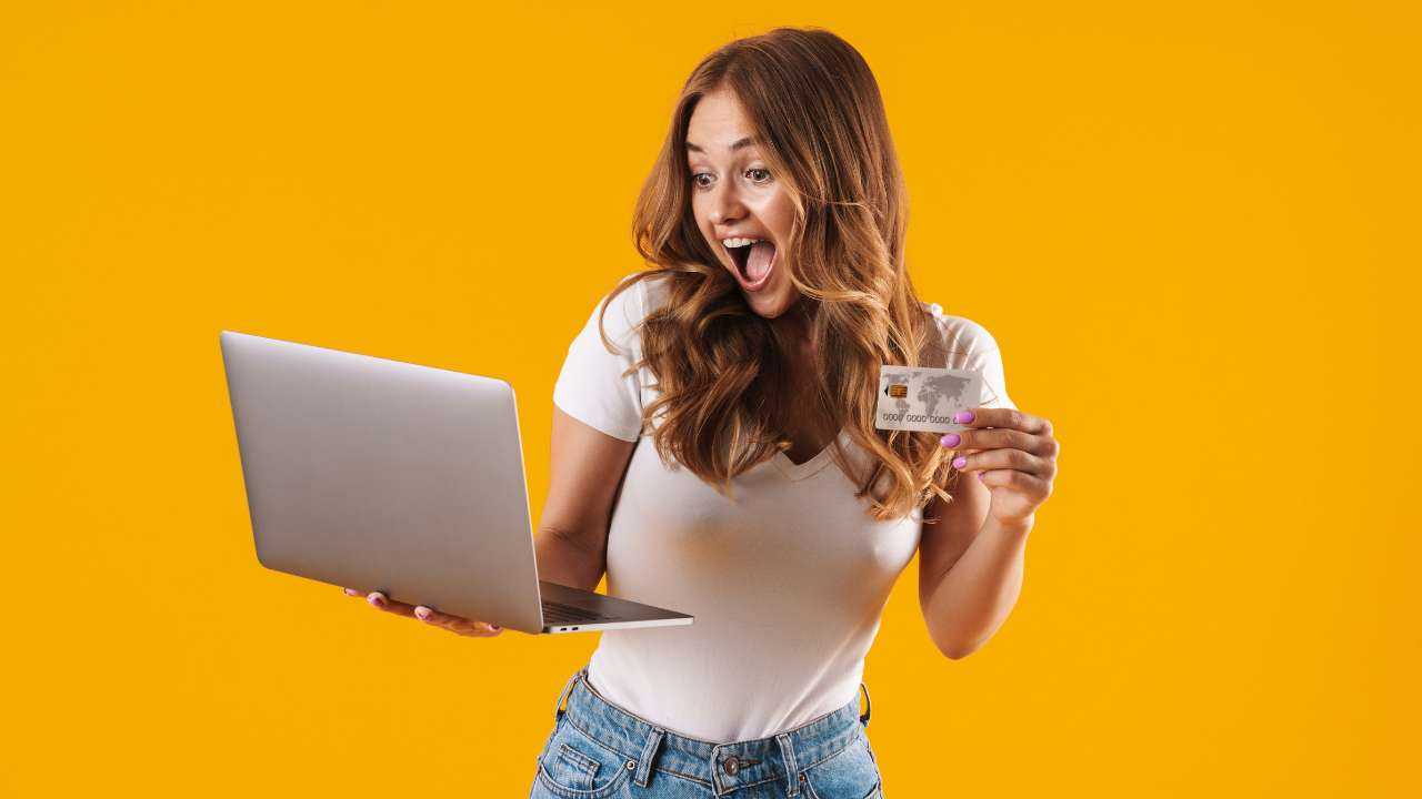 Удивленная женщина с кредитной картой, на которую получен моментальный кредит на карту онлайн, и ноутбуком на желтом фоне