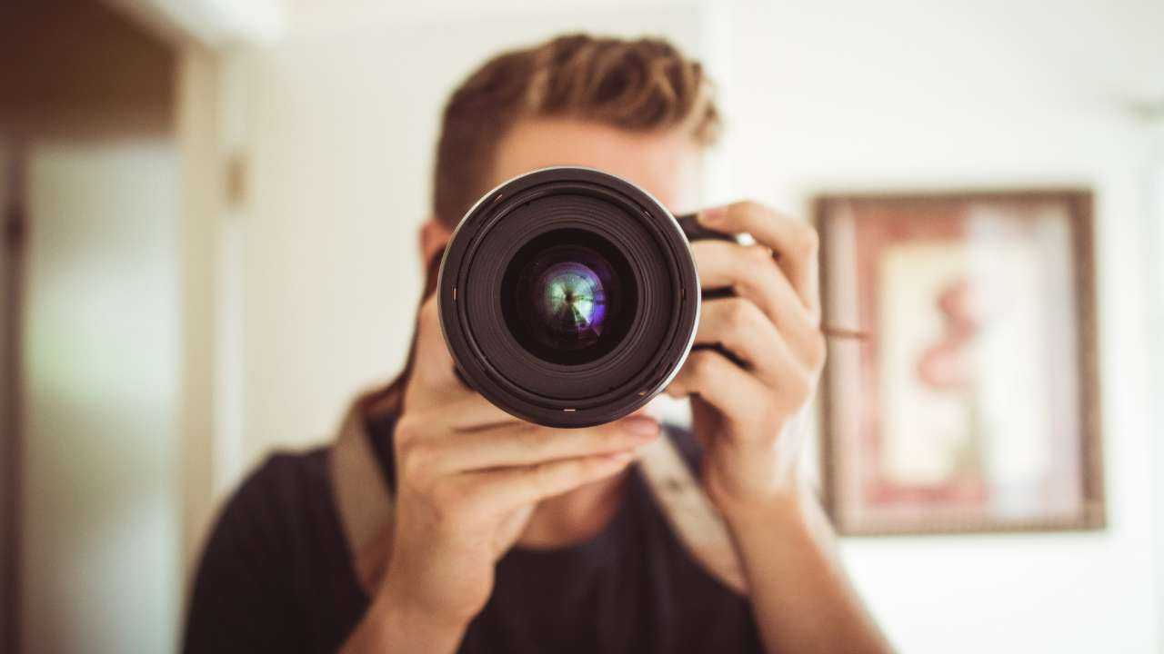 Мужчина монетизирует хобби, держа зеркальную фотокамеру, объектив которой направлен прямо на зрителя, скрывая лицо фотографа