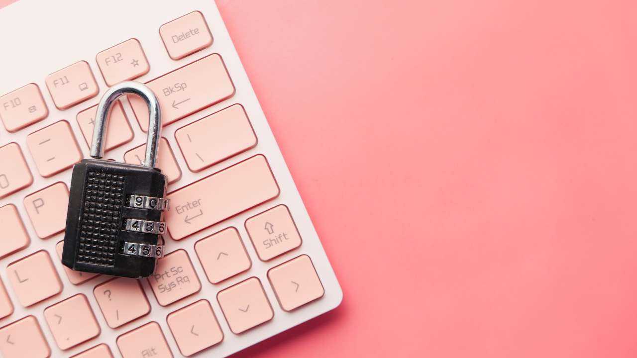 На розовом фоне клавиатура, на которой лежит кодовый замок – необходимости защиты денег от мошенников в сети